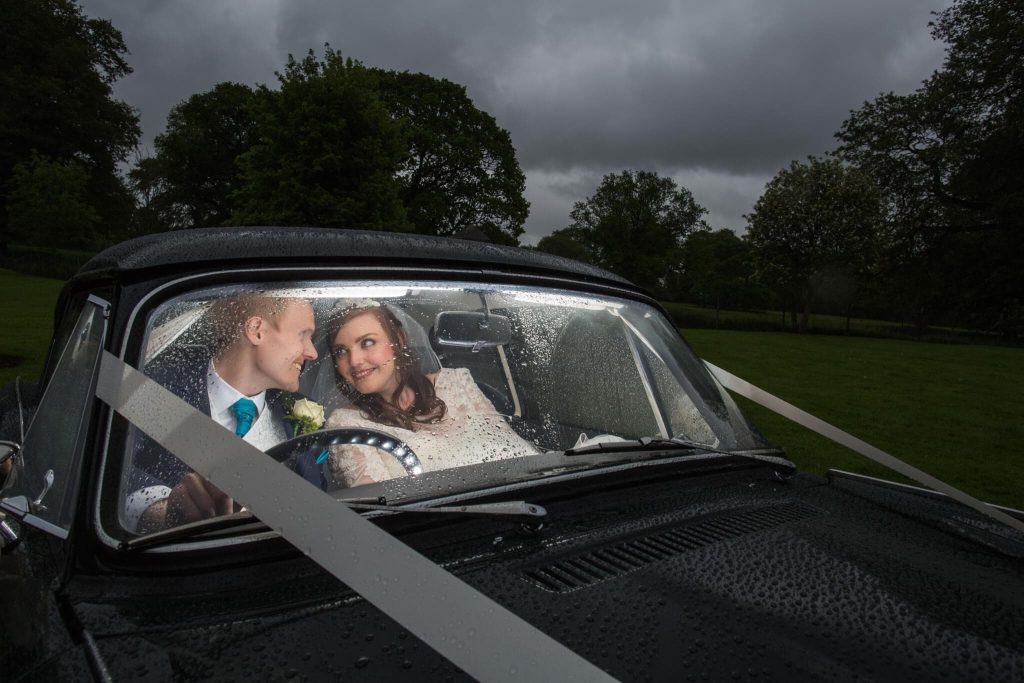 A newlywed couple sits happily inside a rain-covered classic car at their Rothley Court Hotel wedding in Leicestershire. The groom, in a suit with a teal tie, and the bride, in her white dress, smile at each other, surrounded by the lush green landscape.