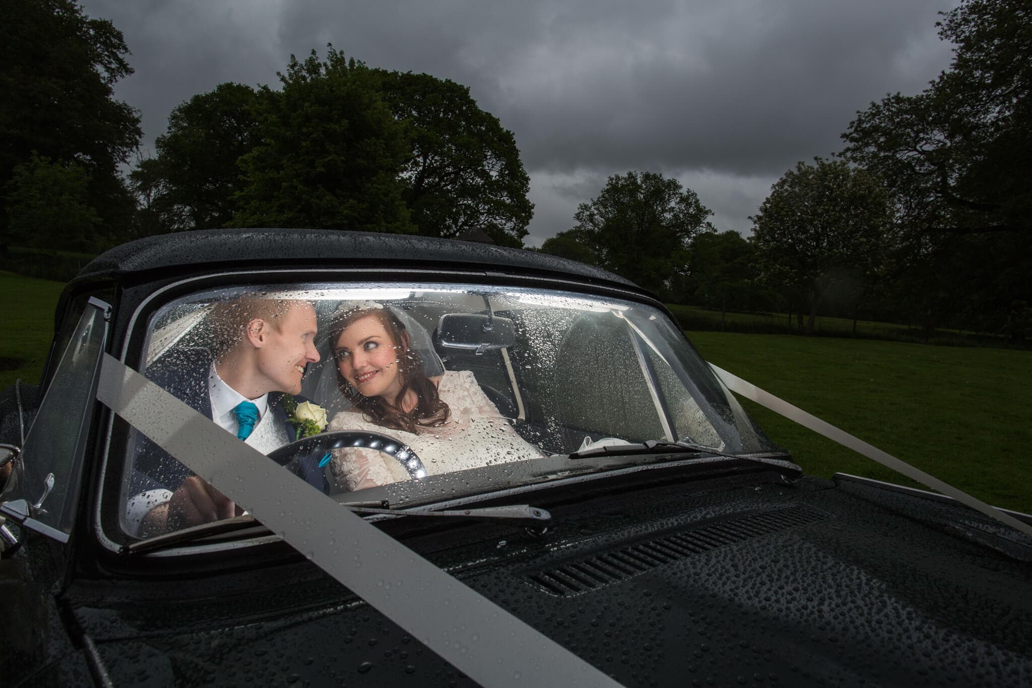 A newlywed couple sits happily inside a rain-covered classic car at their Rothley Court Hotel wedding in Leicestershire. The groom, in a suit with a teal tie, and the bride, in her white dress, smile at each other, surrounded by the lush green landscape.