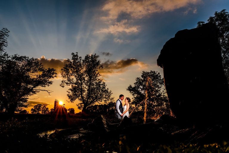 A couple embraces at sunset in Shearsby Bath, Leicestershire. The sun casts rays through clouds, silhouetting trees and a fountain water feature. The sky is a vibrant mix of orange and blue, creating a romantic atmosphere.