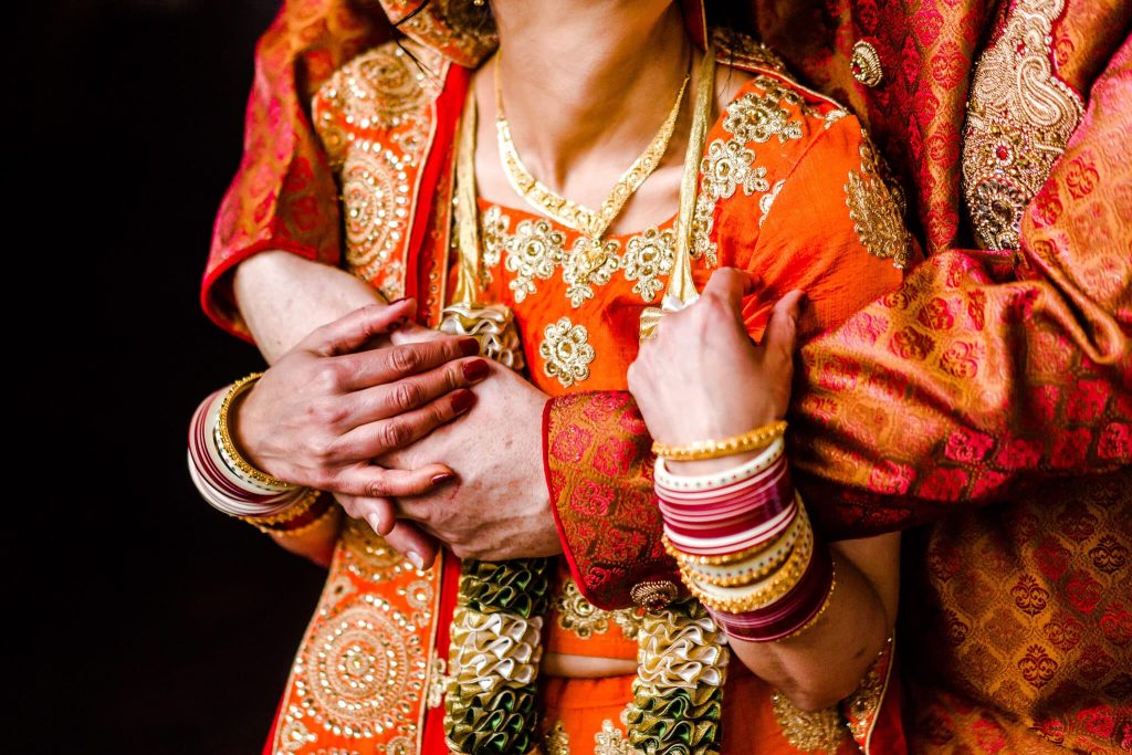 A close-up of a couple at an Indian wedding in colourful traditional attire embracing. They wear vibrant outfits adorned with gold embroidery and intricate patterns. Their hands are interlocked, displaying decorated bangles and jewellery, symbolising a cultural celebration. Taken at a hindu wedding in Nottingham