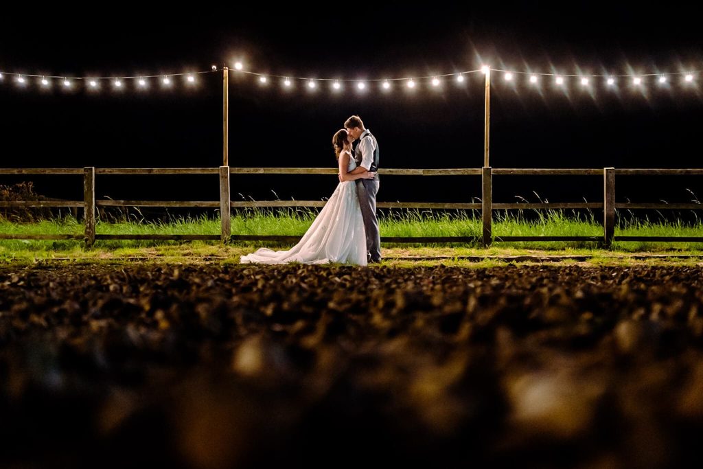 A couple embraces under string lights at night in a field with a wooden fence, capturing the romance of a Swancar Farm Country House wedding. The bride wears a flowing white gown, and the groom dons a white shirt and gray pants. The ground is out of focus, highlighting their intimate scene.