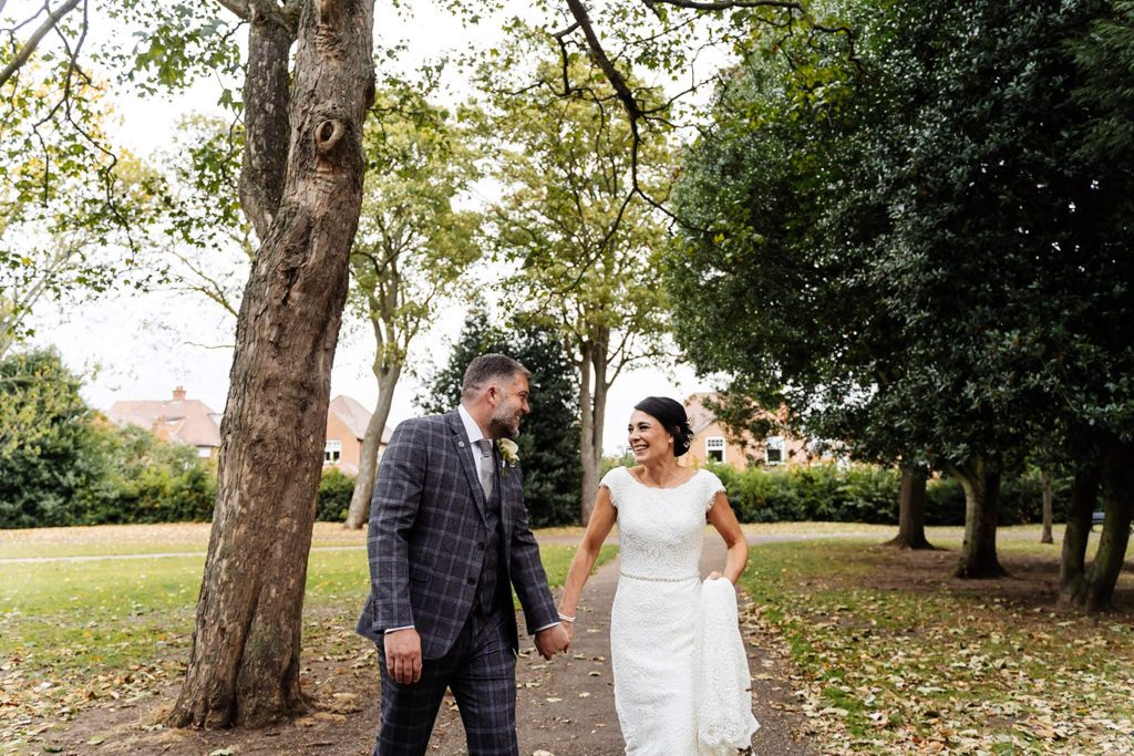 A couple, dressed in wedding attire, joyfully walks hand in hand along a tree-lined path at West Bridgford Park. The groom wears a plaid suit, and the bride is in a white gown. They are surrounded by greenery and fallen leaves in this serene park setting.