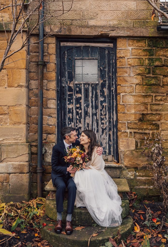 A bride and groom sit on stone steps in front of a rustic wooden door at their charming Bakewell wedding in Derbyshire. The bride holds a bouquet of autumn flowers, and they gaze at each other lovingly as fallen leaves and bare branches complete the picturesque autumn setting.