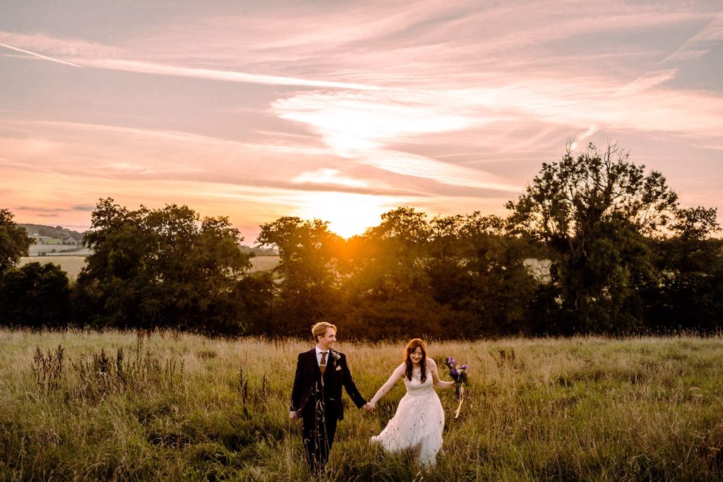 A couple in formal attire walks, during their wedding, hand in hand through a grassy field at sunset near Shottle Hall, Belper. The sky is orange and pink, and trees line the horizon, creating a romantic and serene atmosphere.