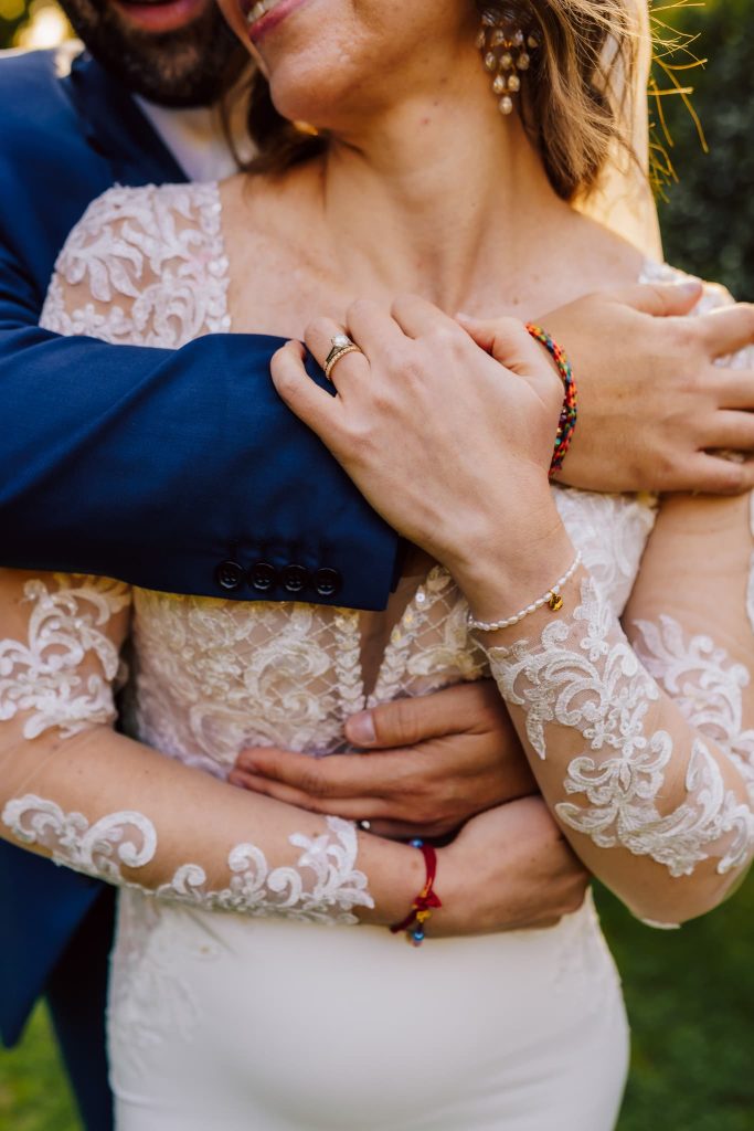 A couple embracing at there Stanwick Hotel wedding, with the man in a blue suit and the woman in a white lace dress. She wears colourful bracelets, and they each have wedding rings. They're outdoors, surrounded by greenery, with sunlight filtering through in a serene fusion of nature and love.