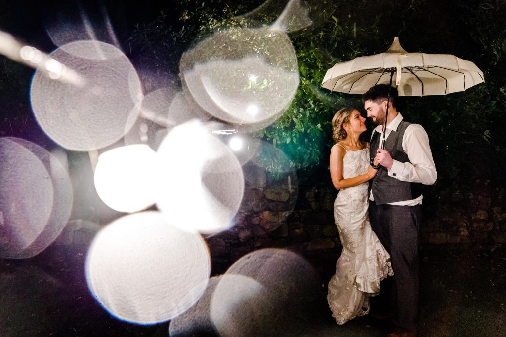 The bride and groom stand under an umbrella, smiling at each other during their rainy wedding day. Outside at night with bokeh light effects surrounding them, the scene captures the romance of Blackbrook House in Derbyshire. The bride's lace dress and the groom's suit add elegance to this magical moment.