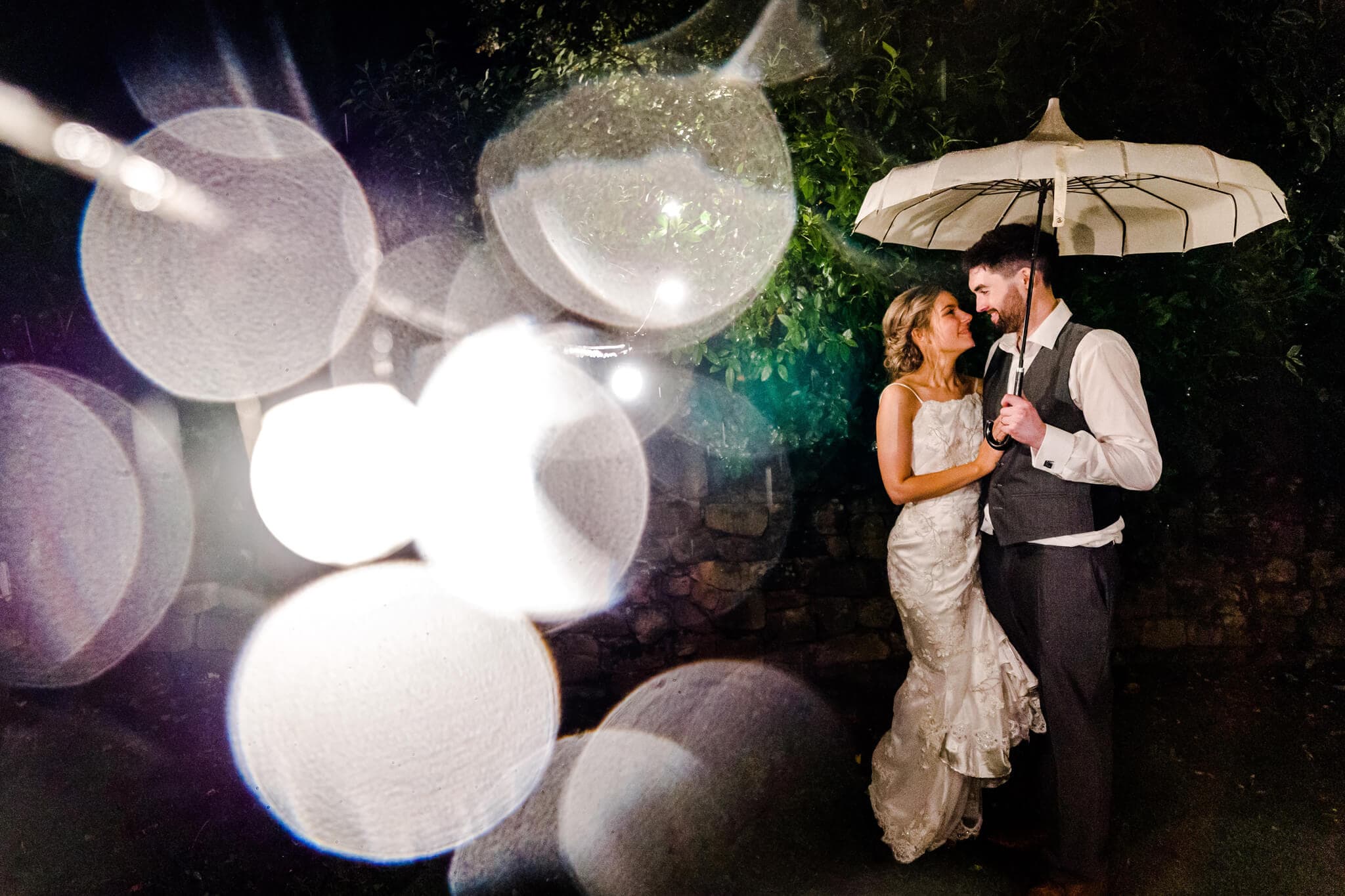 The bride and groom stand under an umbrella, smiling at each other during their rainy wedding day. Outside at night with bokeh light effects surrounding them, the scene captures the romance of Blackbrook House in Derbyshire. The bride's lace dress and the groom's suit add elegance to this magical moment.