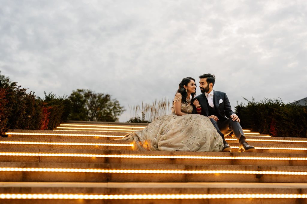 A couple sits on illuminated steps under a cloudy sky at Fairmont Windsor Park. The woman, in an embellished gown, and the man, in a suit, share a loving smile amidst lush greenery—a perfect moment from their Sikh wedding celebration.