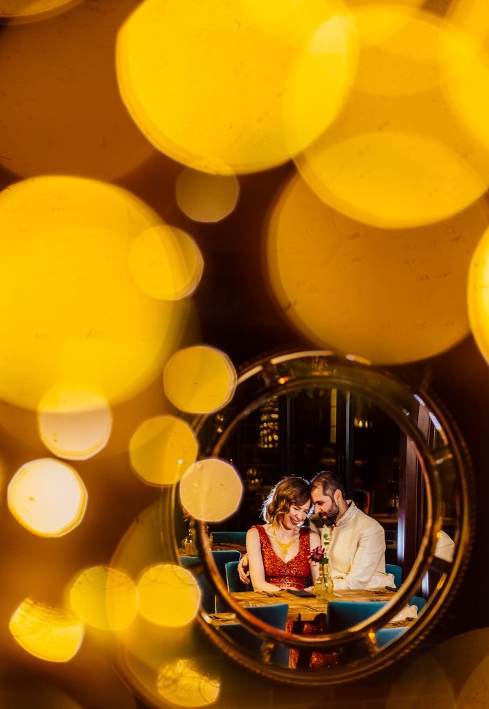 A couple sits closely at a table, smiling and holding hands. Framed by a circular mirror during their night time wedding photos with warm, glowing bokeh lights, they create an intimate and romantic atmosphere.