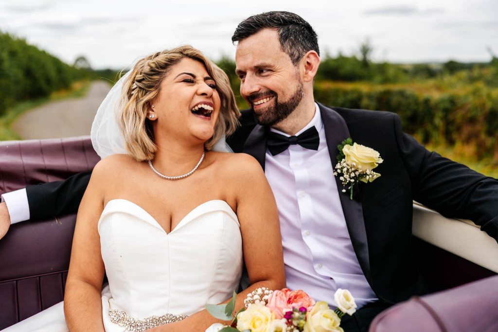A bride and groom laugh joyfully in a classic car at their Blackwell Grange wedding. The bride, in a strapless white gown, holds a bouquet of roses, while the groom wears a black tuxedo with a white rose boutonniere. A scenic road unfolds in the background.