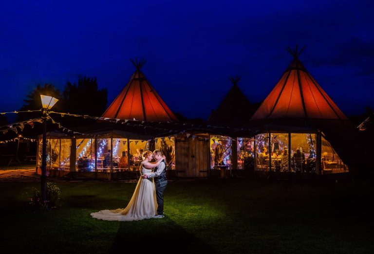 A couple embraces under a vibrant night sky, standing before warmly lit teepee tents decorated with string lights at the picturesque Bridge House Barn in Leicestershire. The scene exudes a romantic and festive atmosphere, perfect for a tipi wedding celebration.