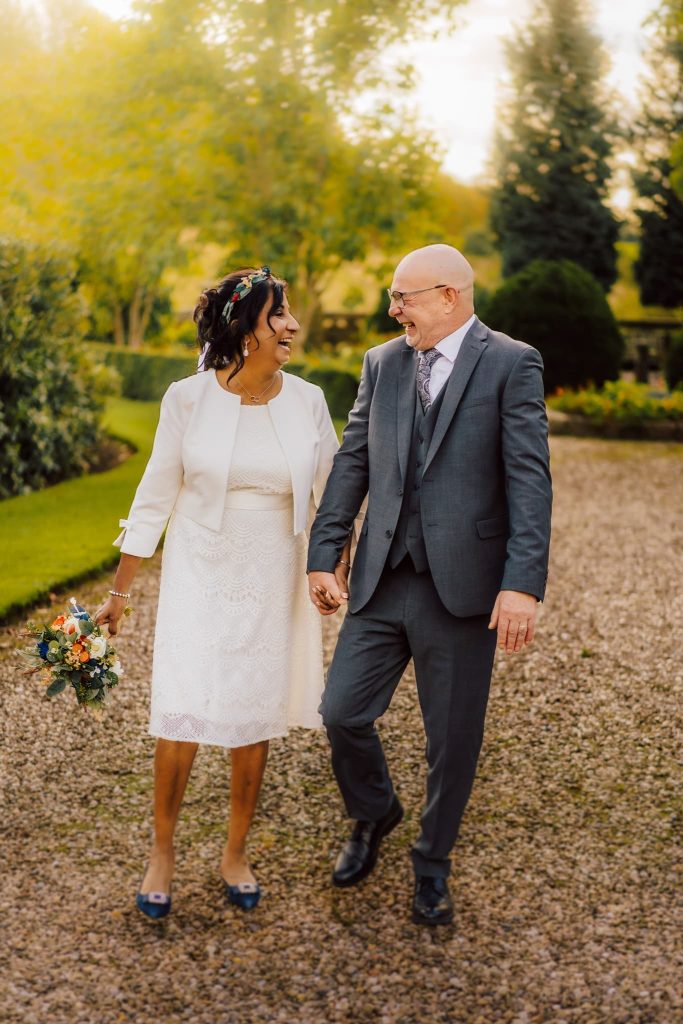 A smiling couple walks hand in hand on a gravel path surrounded by greenery at Beaumanor Hall near Loughborough. The woman, in a white dress and jacket, holds a bouquet, while the man sports a gray suit. They appear joyful, savouring the sunny day outdoors.