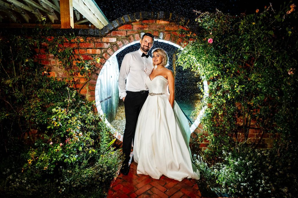 A bride and groom stand smiling under a circular archway at Blackwell Grange, surrounded by greenery and flowers. It is nighttime, and they're illuminated by soft lighting. The bride wears a white gown, the groom in a white shirt and black trousers, while raindrops add magic to the scene.