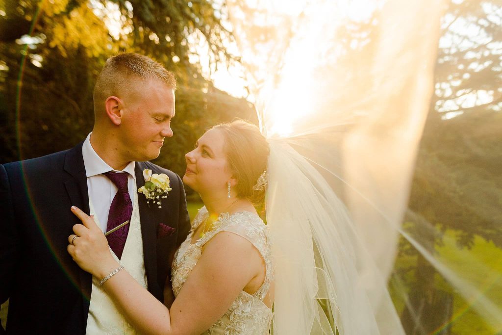 A bride and groom gaze into each other's eyes, smiling warmly, at Prestwold Hall. The bride's veil is gently blown by the breeze as they stand bathed in sunlight filtering through trees, creating a golden glow reminiscent of a sunset photo.