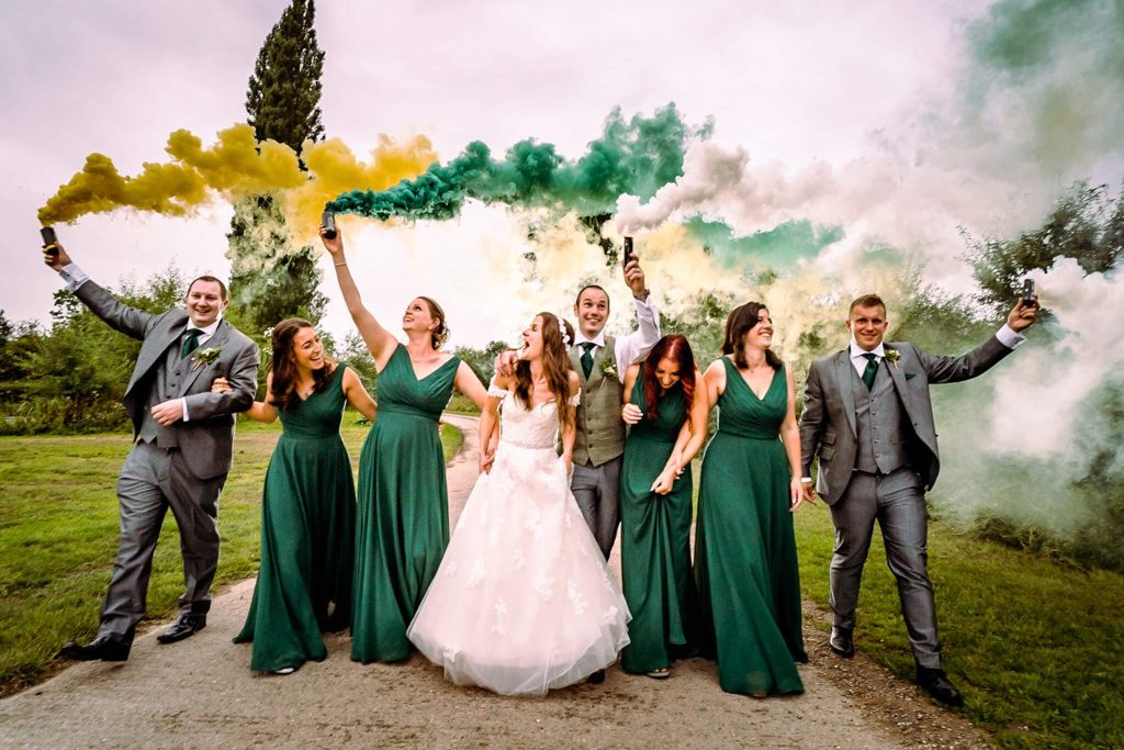 A bride and groom, surrounded by their bridal party at Doddington Hall in Lincolnshire, walk outdoors. The bridesmaids wear green dresses, and the groomsmen are in grey suits. They hold colourful smoke flares, creating a vibrant background—a perfect example of alternative wedding photography amidst lush greenery.