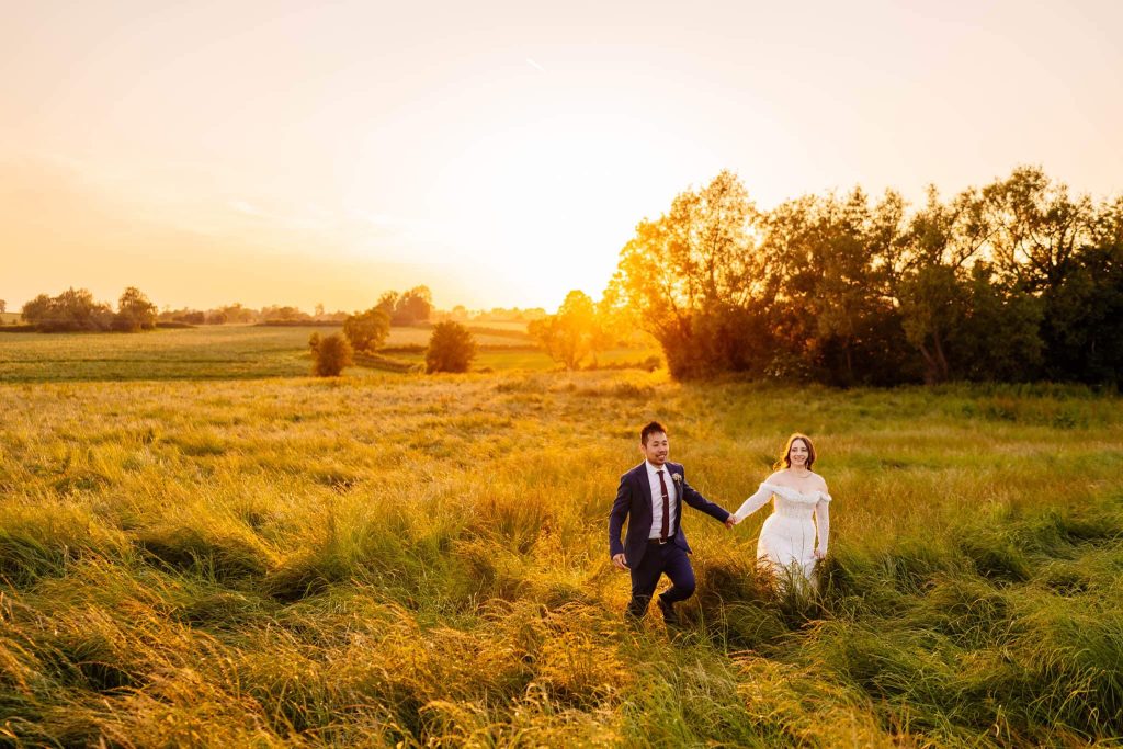 A couple holding hands walks through a grassy field at sunset at Grangefields, Derbyshire. The man is in a suit, and the woman is in a white dress. Sunset photos capture the sun as it sets, casting a warm glow over the serene landscape.