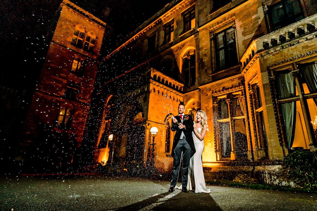 Outside the grand, illuminated Walton Hall in Warwickshire, a couple celebrates their wedding at night. The man is popping a champagne bottle as the woman smiles beside him. A champagne spray fills the air, adding a festive atmosphere to the elegant scene.