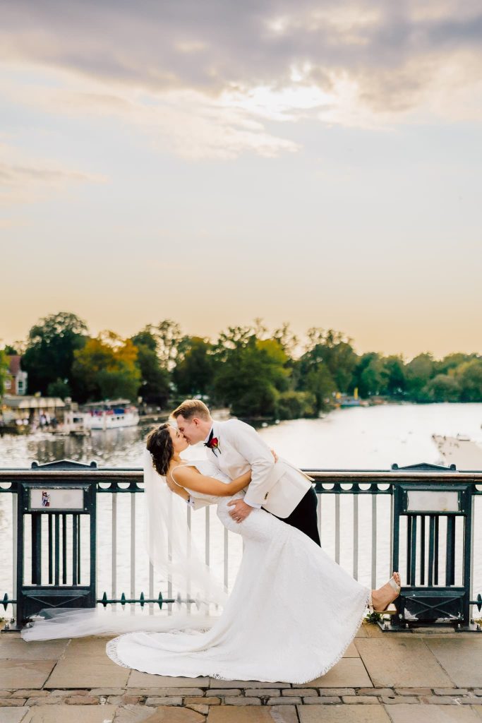 A couple in wedding attire shares a romantic dip and kiss on a bridge overlooking the River Thames at sunset. The groom wears a light suit, and the bride dazzles in a white gown with a veil. Boats and trees line the river near Windsor, adding to their enchanting Eton wedding backdrop.