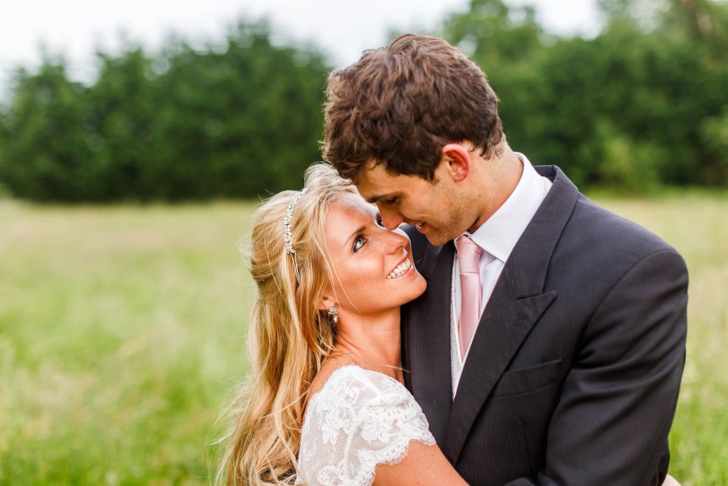 A bride and groom embrace in a grassy field. The bride, wearing a white dress and tiara, gazes up smiling at the groom, who is wearing a dark suit and light pink tie. Green trees are visible in the background.