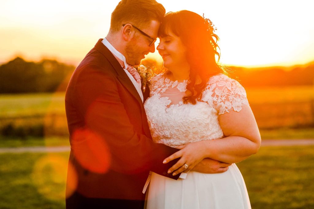 A couple embraces lovingly at sunset on the historic Bosworth Battlefields in Leicestershire. The sun casts a warm glow over them as the bride, in a white lace dress, and the groom, in a suit, stand outdoors on the grassy field, smiling at each other.