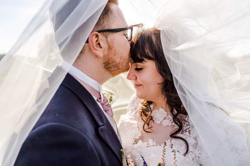 A bride and groom share an intimate moment under a white veil at Bosworth Battlefields. The groom, in a dark suit, gently kisses the bride’s forehead. The bride, in a lace gown, smiles softly with her eyes closed, surrounded by soft natural light in picturesque Leicestershire.