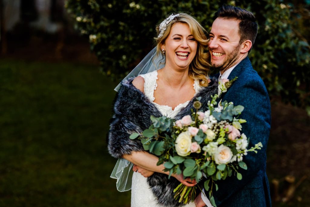 A joyful couple smiles on their wedding day at Shottle Hall. The bride, in a white dress and holding a bouquet of flowers, stands beside the groom in a blue suit. They appear happy and relaxed, surrounded by the serene greenery characteristic of this winter wedding setting.
