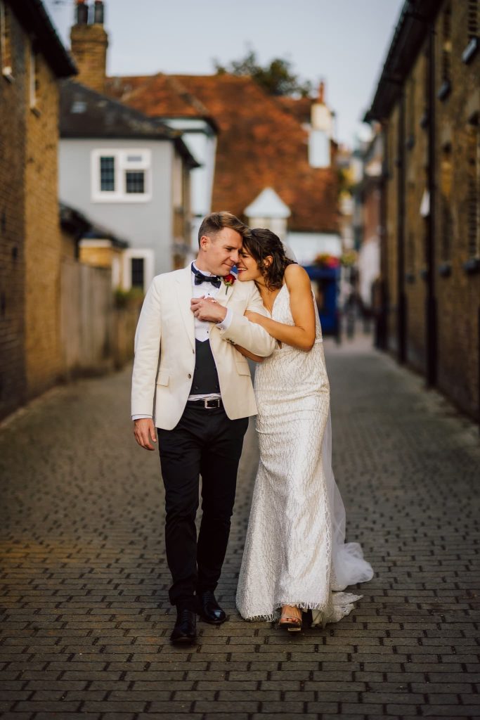 A couple dressed in wedding attire strolls down a cobblestone street during their Windsor wedding The woman in a white gown leans affectionately on the man in a white jacket and black pants, surrounded by charming brick buildings under a blue sky.