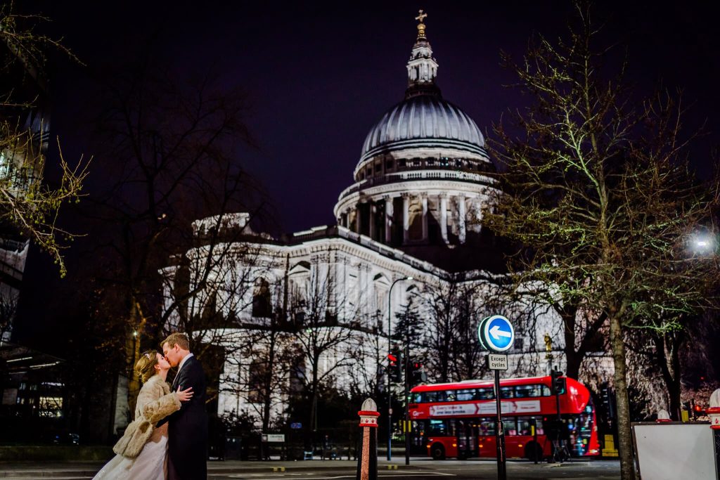 A couple embraces and kisses at night in front of an illuminated St. Pauls Cathedral, creating a romantic London wedding scene as a red double-decker bus glides past. Silhouetted trees add to the atmospheric backdrop.
