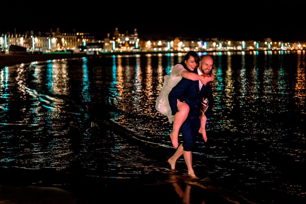 A bride in a white dress playfully piggybacks on the groom, who is barefoot in a dark suit, walking in the Dorset sea at night. City lights reflect on the calm water behind them, creating a romantic and joyful Weymouth scene that perfectly captures their DIY wedding spirit.