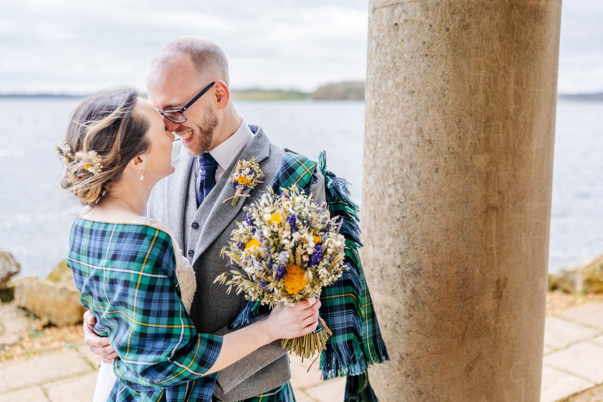 A couple embraces near a stone column by the water at Normanton Church, their tartan clothing reflecting the joyous spirit of their Rutland Water wedding. The woman cradles a bouquet of flowers as they savor this warm, happy moment together.