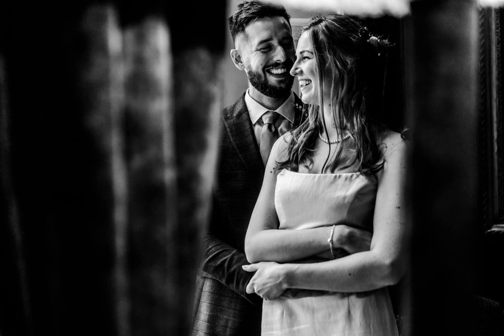 A black and white photo captures a couple sharing a joyful moment indoors at Prestwold Hall. The man, in a suit, stands behind the woman in a simple dress, holding her with a warm smile. Both appear to be looking at each other happily despite the rainy day wedding outside.