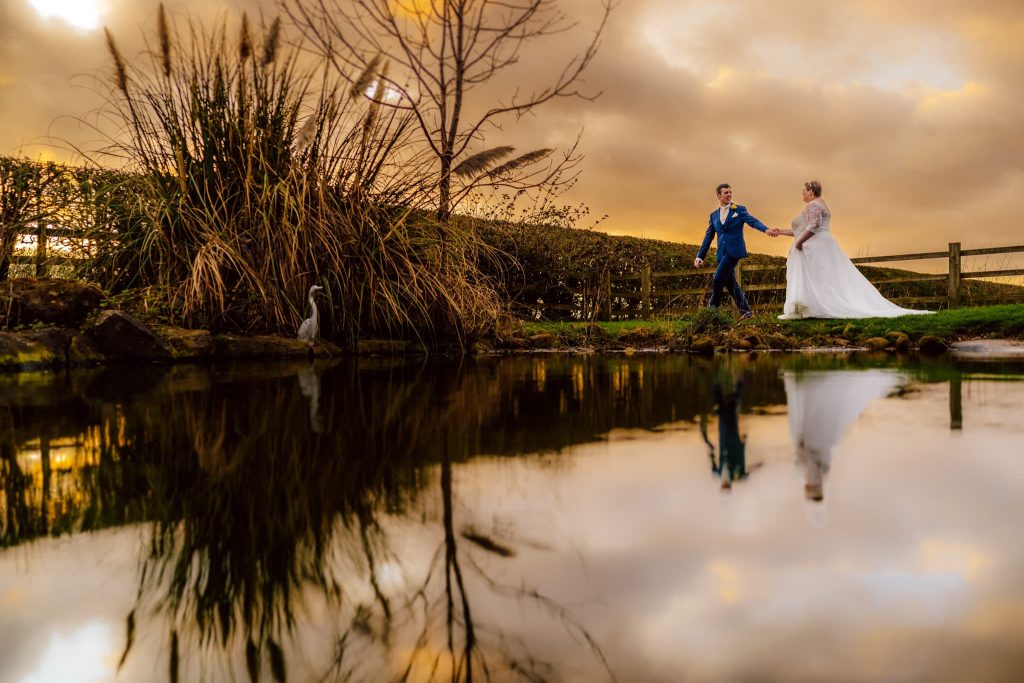 A bride and groom walk hand in hand by the tranquil pond at Fishlake Mill, Lincolnshire, as the sunset paints the sky orange. Clouds reflect in the water while tall grasses and a heron stand by its edge, crafting a serene and romantic scene.