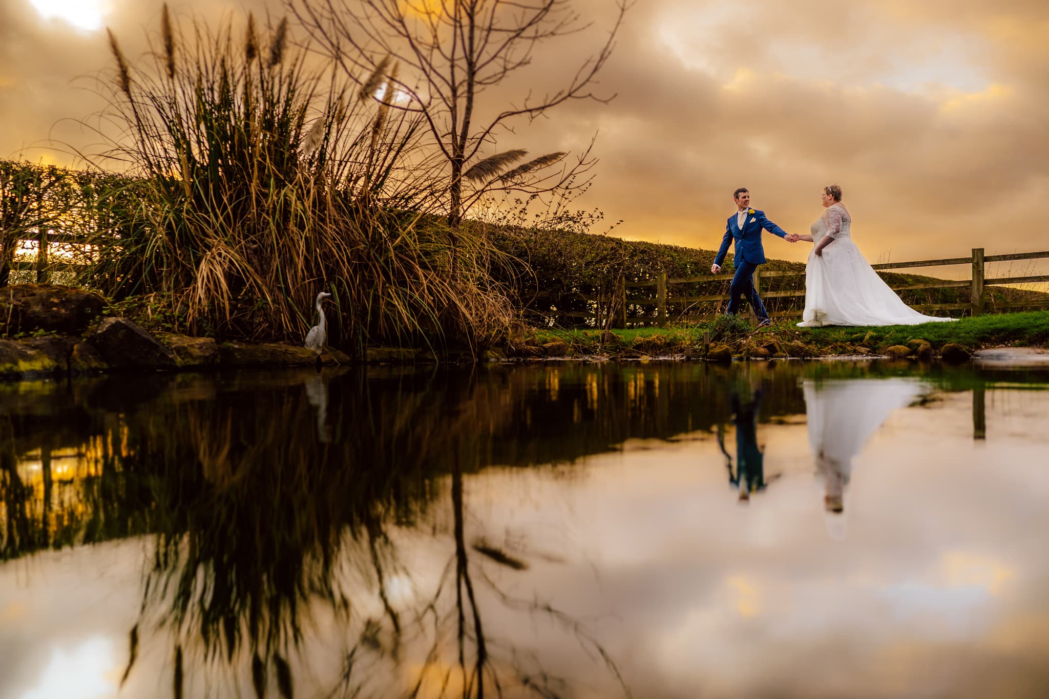 A bride and groom walk hand in hand by the tranquil pond at Fishlake Mill, Lincolnshire, as the sunset paints the sky orange. Clouds reflect in the water while tall grasses and a heron stand by its edge, crafting a serene and romantic scene.