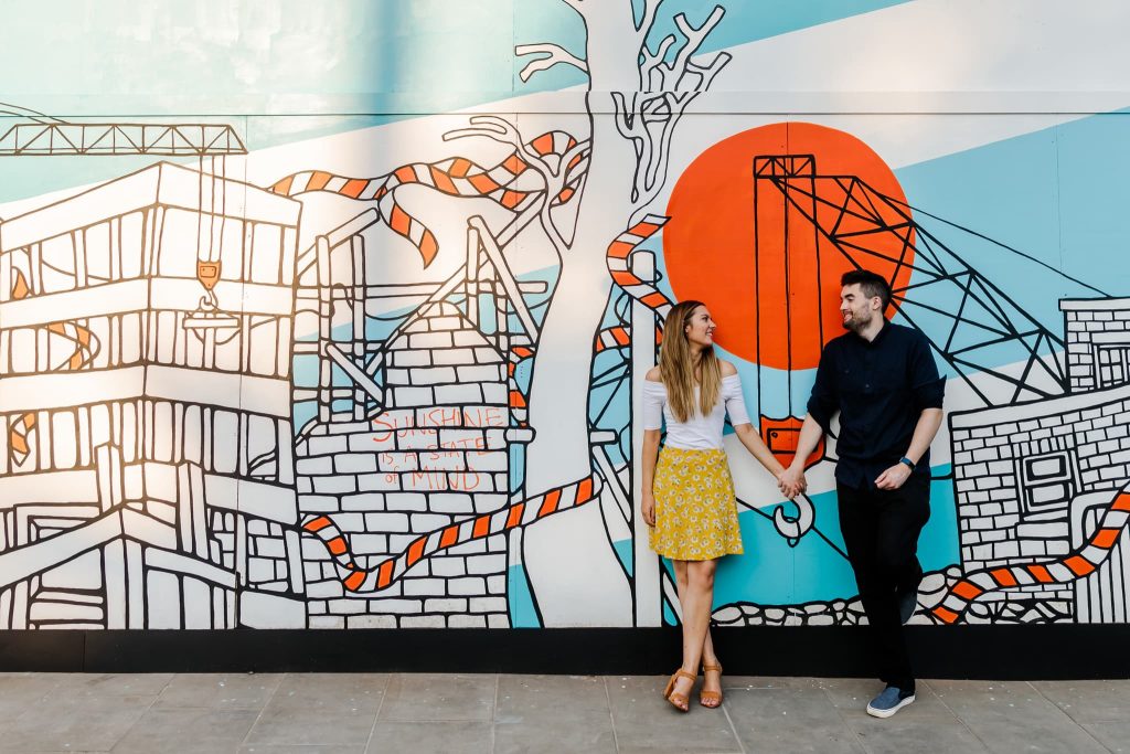 A man and woman hold hands in front of a vibrant mural featuring industrial and abstract elements, capturing the spirit of a Sheffield city pre-wedding scene. The man wears dark clothing, and the woman sports a white top with a yellow skirt, both smiling warmly at each other.