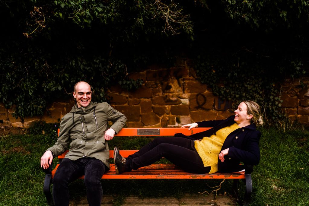 Two people sit on a red bench in a park, smiling and laughing as if capturing a perfect pre-wedding moment in Warwickshire. One wears a green jacket, the other a yellow sweater. Lush greenery surrounds them, adding natures warmth to the scene.