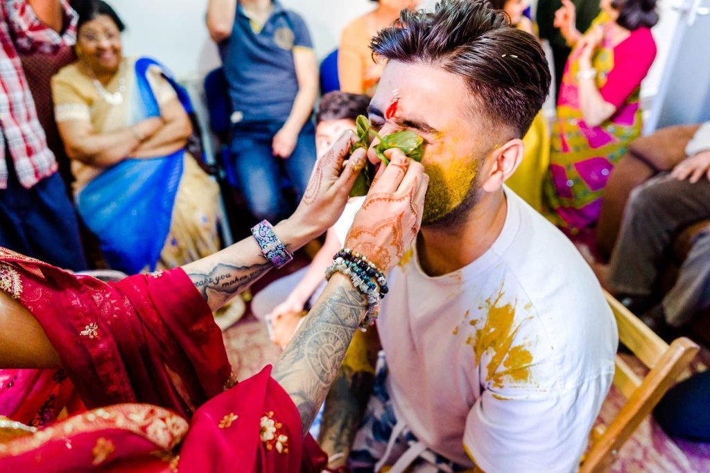 During an Indian traditional pre-wedding celebration, a man in a white t-shirt sits on a chair, his face covered with yellow powder. A person in a red sari applies more powder as people in vibrant attire watch the joyful ritual with smiles.