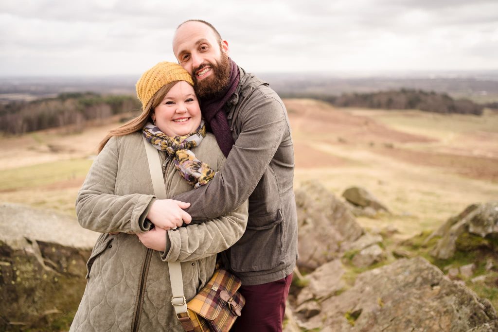 A couple is smiling and embracing on a rocky hillside with the scenic landscape of Bradgate Park in the background. The woman wears a yellow beanie and scarf, while the man is in a grey jacket. Its a cloudy day with fields and hills stretching into the distance, perfect for pre-wedding memories.