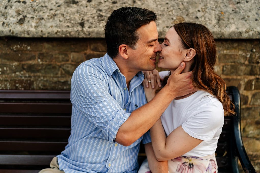 A couple in Oakham sits on a bench, about to kiss. The man, in a blue striped shirt, gently holds the womans face. She wears a white top and floral skirt. They both smile, surrounded by a stone wall in the background, enjoying this tender pre-wedding moment.