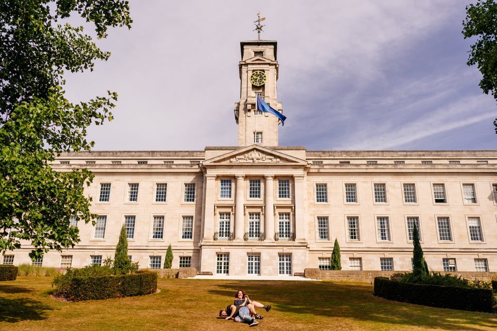 A couple roll down the hill in front of Nottingham University's historic building with a clock tower. The classical architecture, featuring columns and a waving flag, provides a stunning pre-wedding backdrop. The sky is clear with a few clouds, and trees beautifully frame the scene.