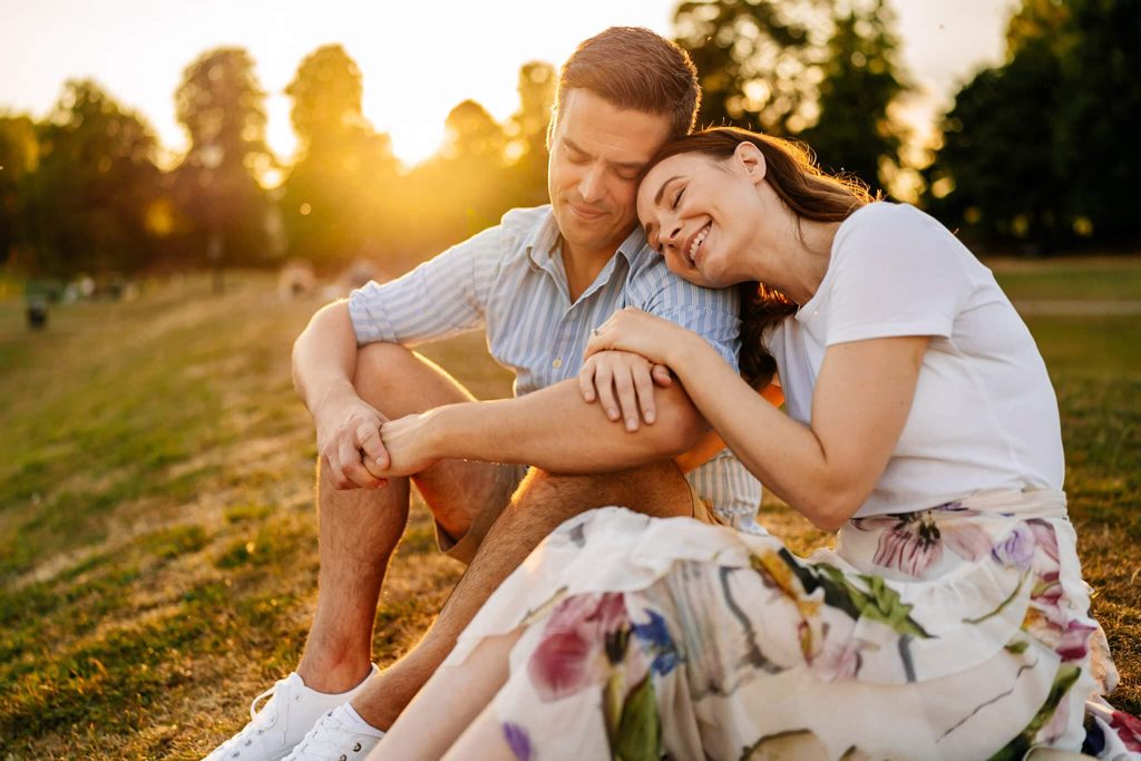 A couple enjoys a serene moment in Oakhams park during sunset, sitting on the grass as part of their pre-wedding shoot. The woman rests her head on the mans shoulder, with trees and a golden sky in the background. They both appear content amidst this picturesque setting.