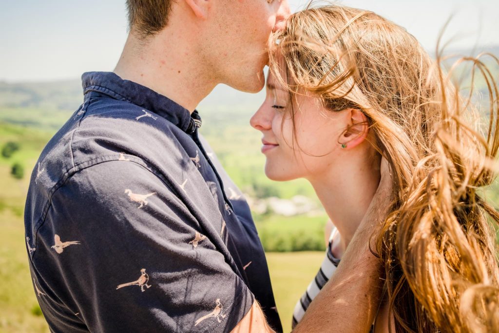 In a tender embrace beneath the sunny skies of the Peak District, a man kisses a womans forehead. Lush green hills form the backdrop as her hair dances in the breeze. Both seem content and peaceful, captured in their perfect pre-wedding moment.