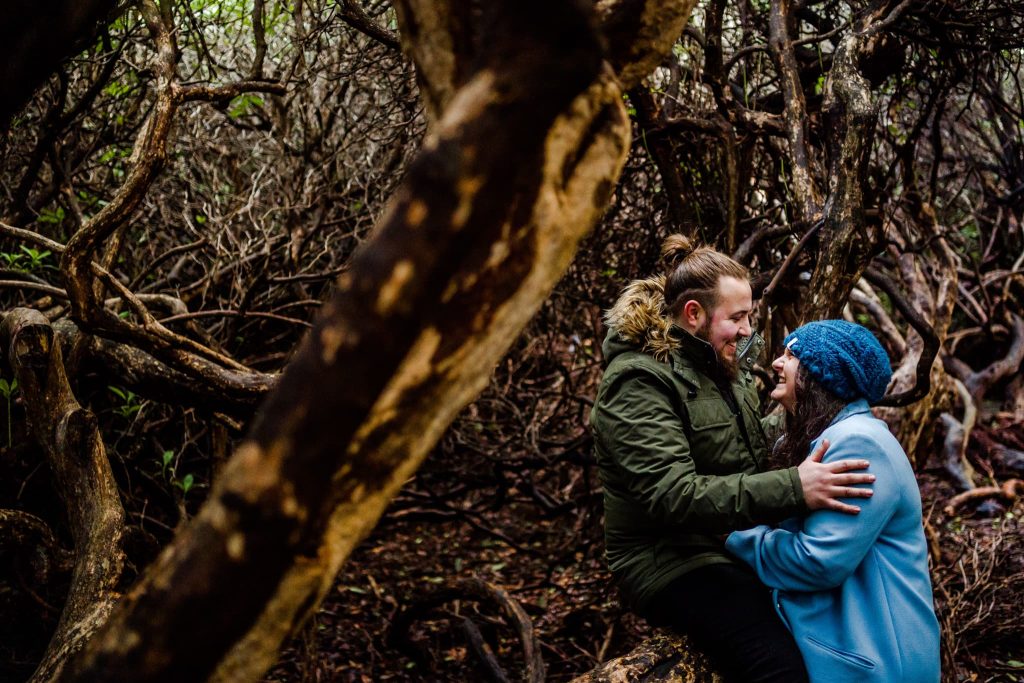 In the enchanting forest of Beacon Hill, Leicestershire, a couple sits among twisted tree branches. They smile at each other; the person on the right dons a blue coat and hat, while their partner wears a green coat with a fur-trimmed hood—a perfect pre-wedding moment captured in natures embrace.