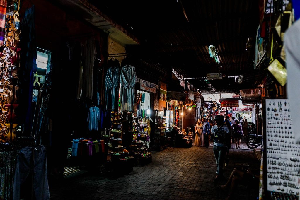 A dimly lit indoor market reminiscent of Marrakesh markets, with stalls offering fabrics, crafts, and jewelry perfect for pre-wedding preparations. Shoppers stroll along the narrow pathway between vibrant displays under a metal roof.