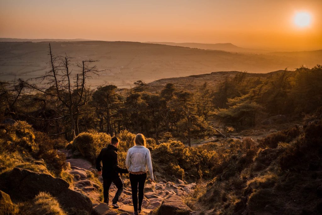 A couple hikes down a rocky trail in the Peak District at sunset, surrounded by trees and a wide landscape. The sky glows orange as the sun sets on the horizon, creating a peaceful and warm pre-wedding atmosphere.