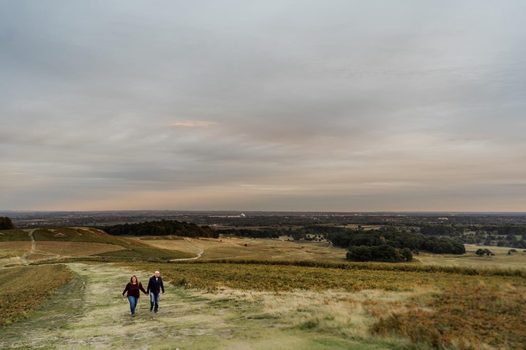 Two people walk along a grassy path in a vast, open landscape under a cloudy sky at Bradgate Park. The backdrop features rolling hills, scattered trees, and a distant horizon, crafting a serene and expansive pre-wedding atmosphere.