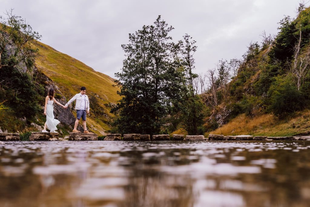A couple holds hands while walking on stepping stones across a shallow stream in Dovedale. They are surrounded by a picturesque landscape with rolling hills, lush greenery, and a cloudy sky in the background, creating the perfect pre-wedding scene.