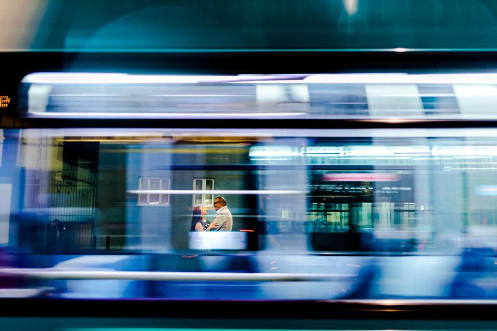 A motion-blurred image of a fast-moving train captures two people sitting inside, facing each other, perhaps sharing dreams in a Leicester city pre-wedding moment. The trains exterior gleams with blue and white hues, reflecting the cityscape and distant buildings visible in the background.