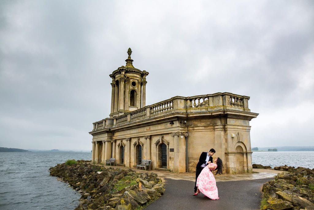 A couple poses romantically in front of a historic stone building by Rutland Water. The overcast sky adds a dramatic backdrop to the pre-wedding scene, where the woman wears a pink dress and the man is in a dark suit.