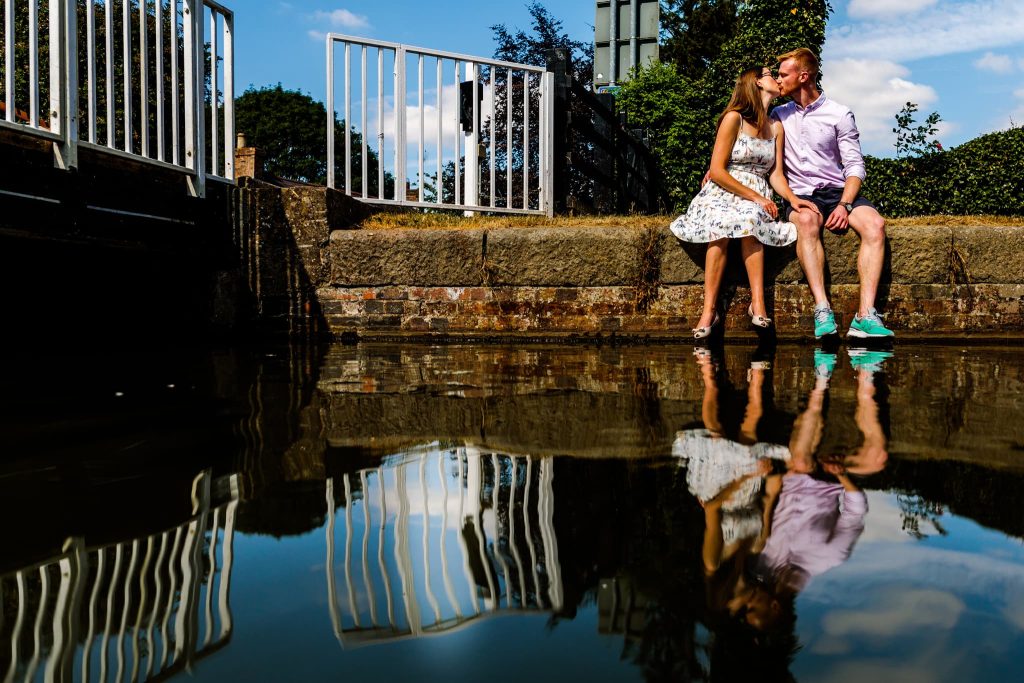 A couple sits by a canal in Leicestershire, sharing a pre-wedding romantic moment. They are seated on a stone ledge, with their reflection visible in the calm water below. The scene is framed by a bright, sunny day with blue skies and green foliage.