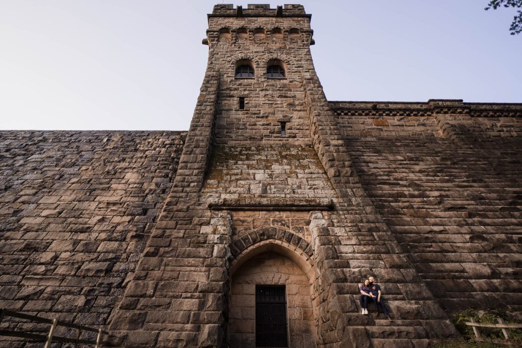 A couple sit on the steps of a large, ancient stone building with a tall tower, reminiscent of the majestic architecture found in the Peak District. The weathered structure features a main arched entrance and various windows. The clear sky enhances this serene pre-wedding setting.